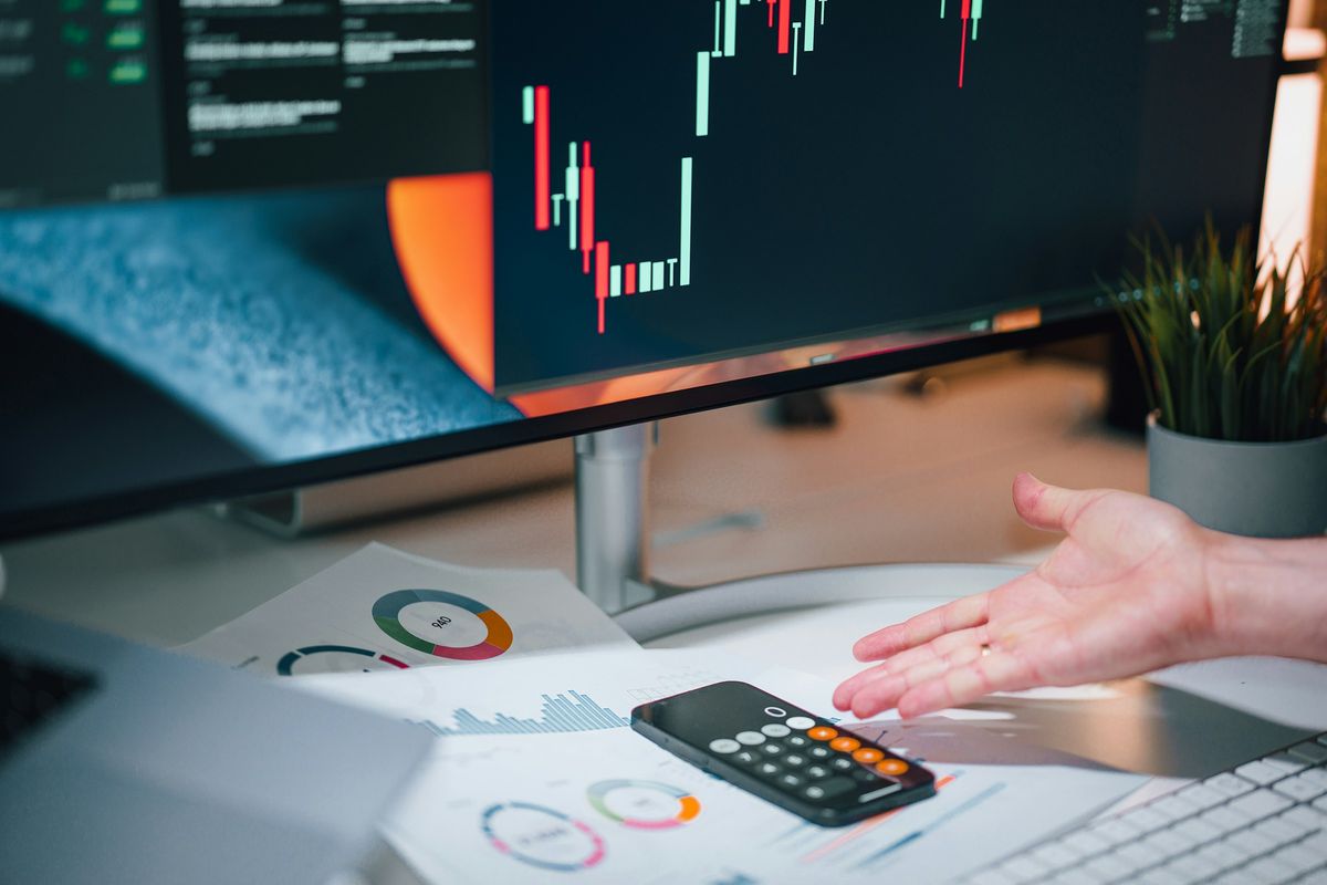 A person's hand reaching over financial charts and reports on a desk, with a computer monitor displaying colorful data analytics in the background and a calculator nearby